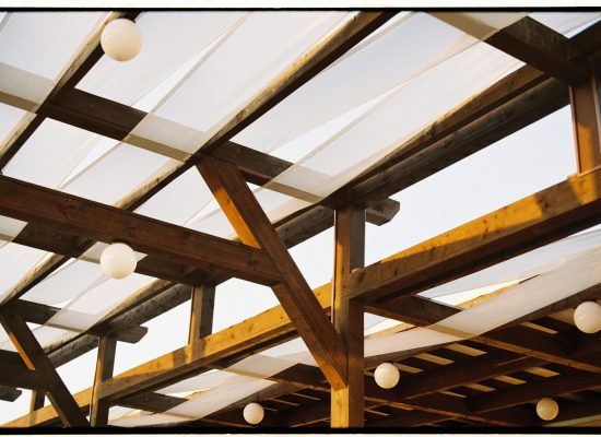 A detailed view of a rustic wooden pergola with white fabric and round light bulbs.